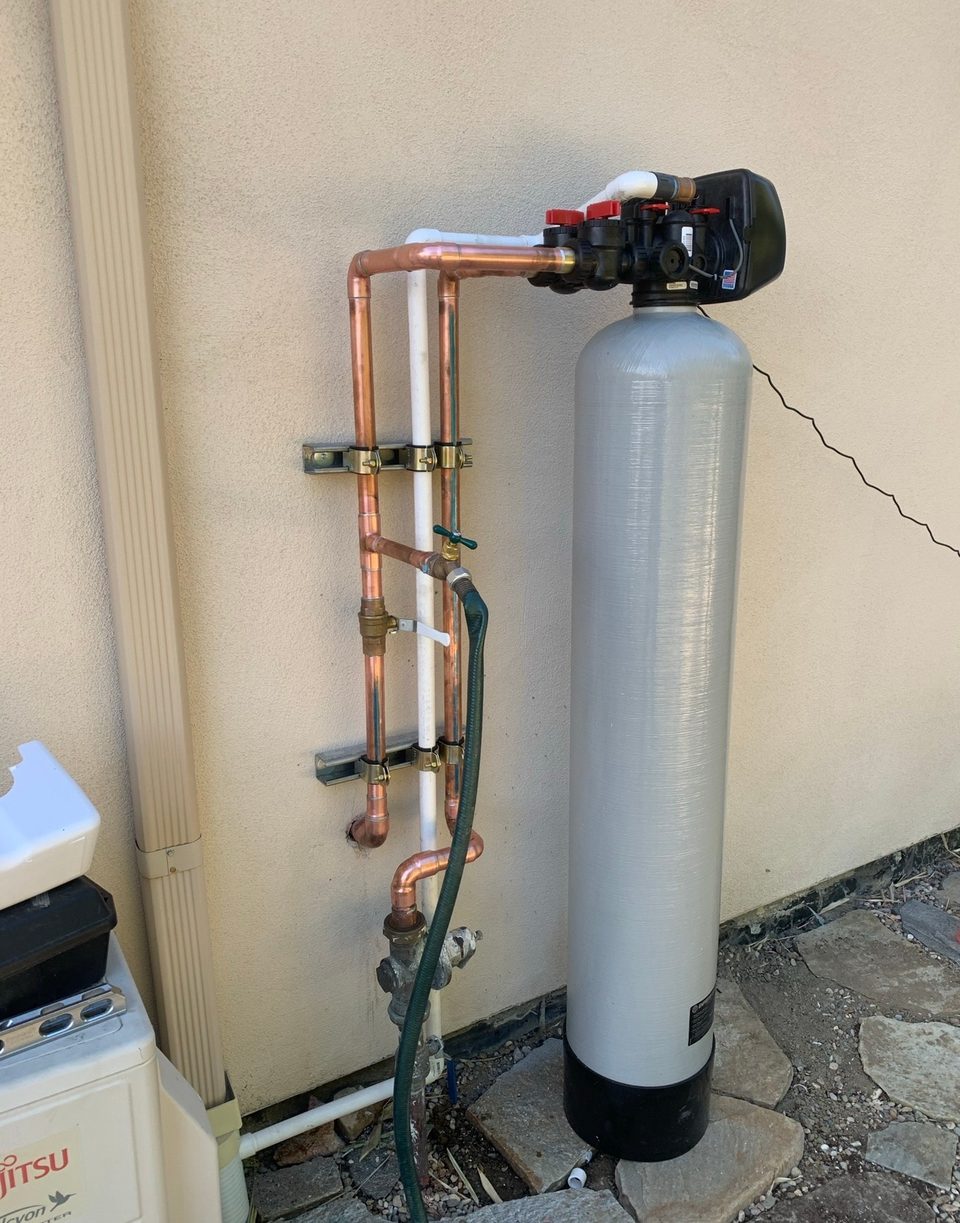 Whole-house water filtration tank connected with copper piping and a bypass valve against a stucco exterior wall