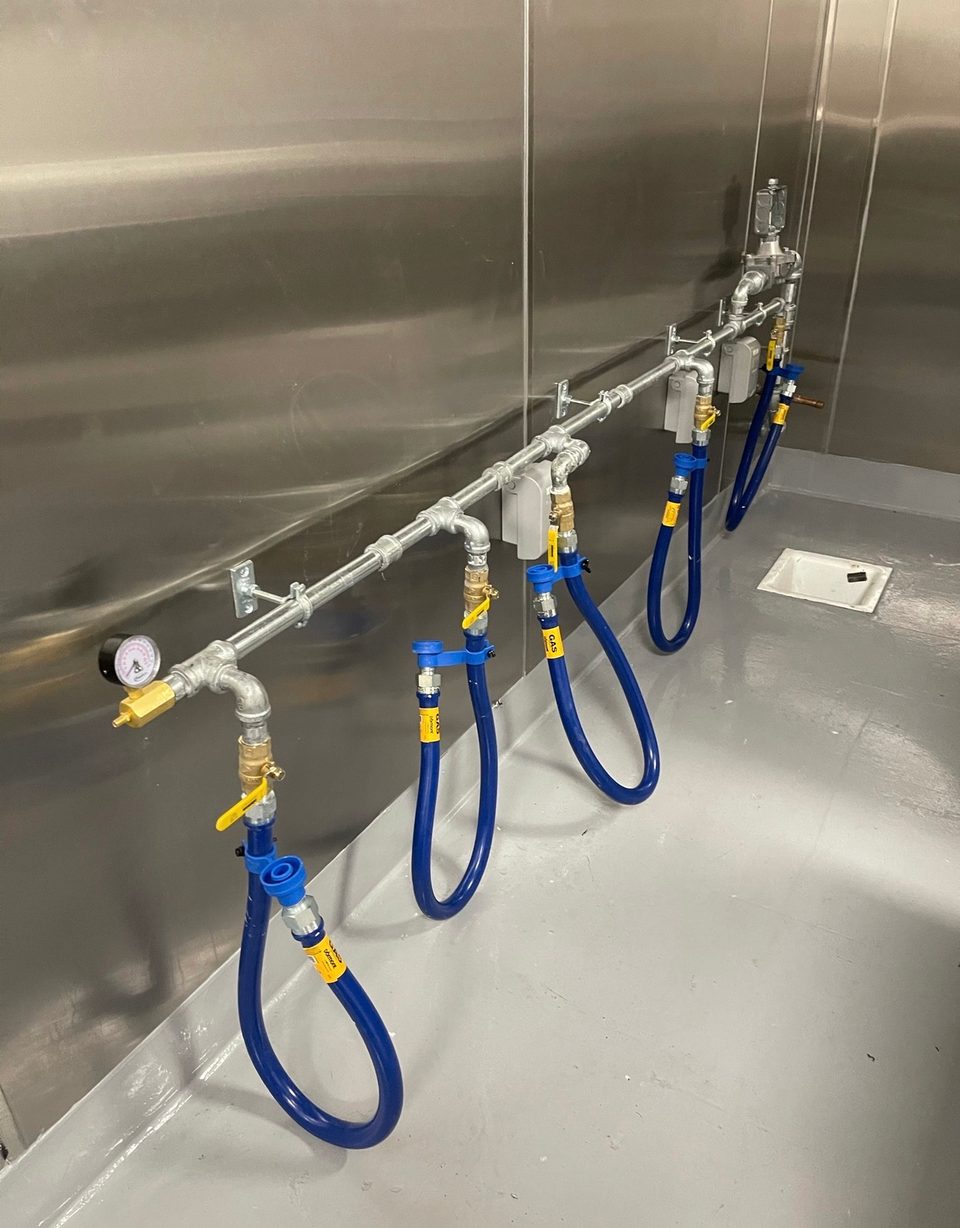 Blue commercial gas connectors and shutoff valves running along a stainless steel commercial kitchen wall