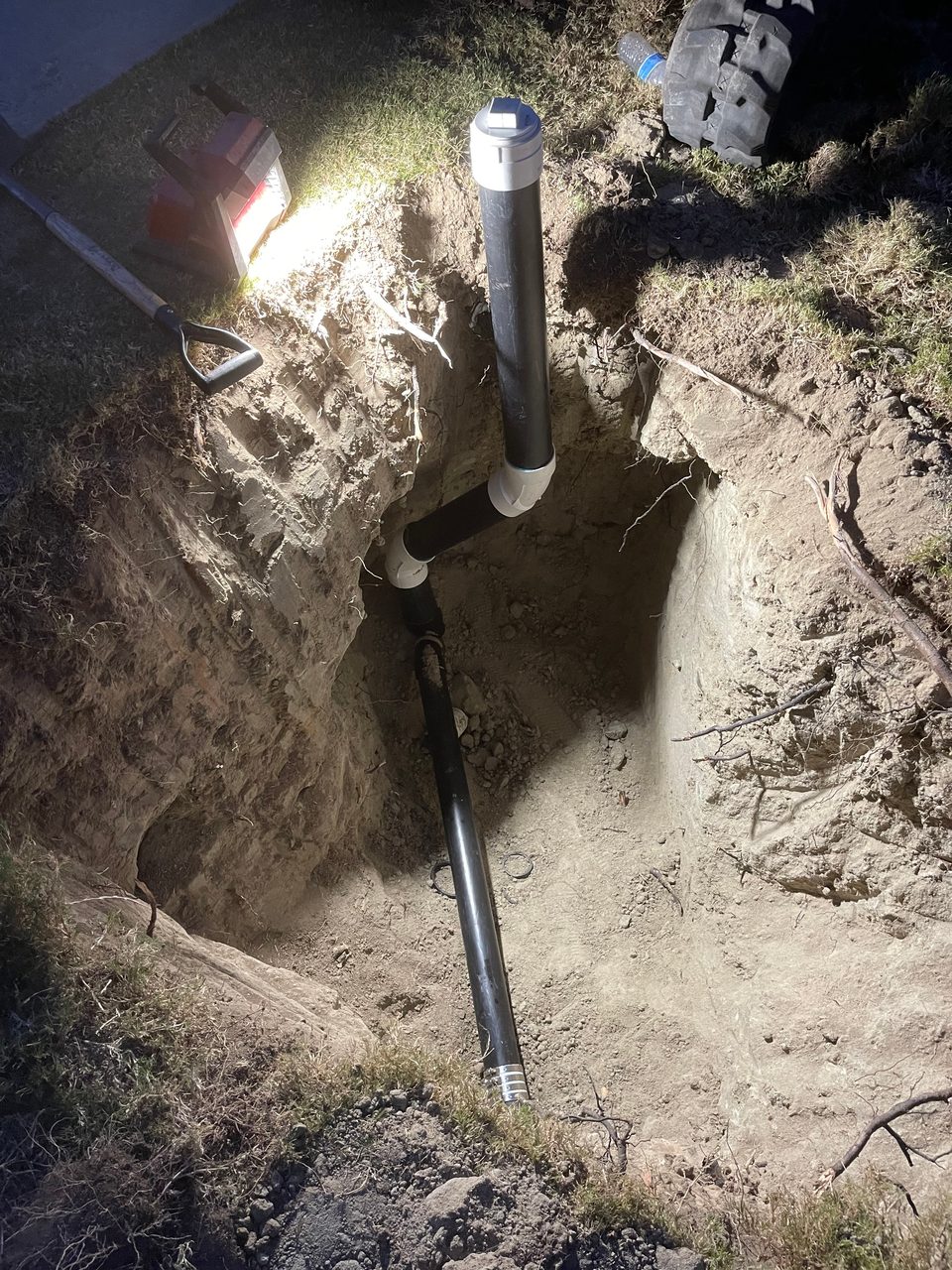 Black ABS sewer line with a white cleanout riser installed in an open trench, photographed at night under work lights