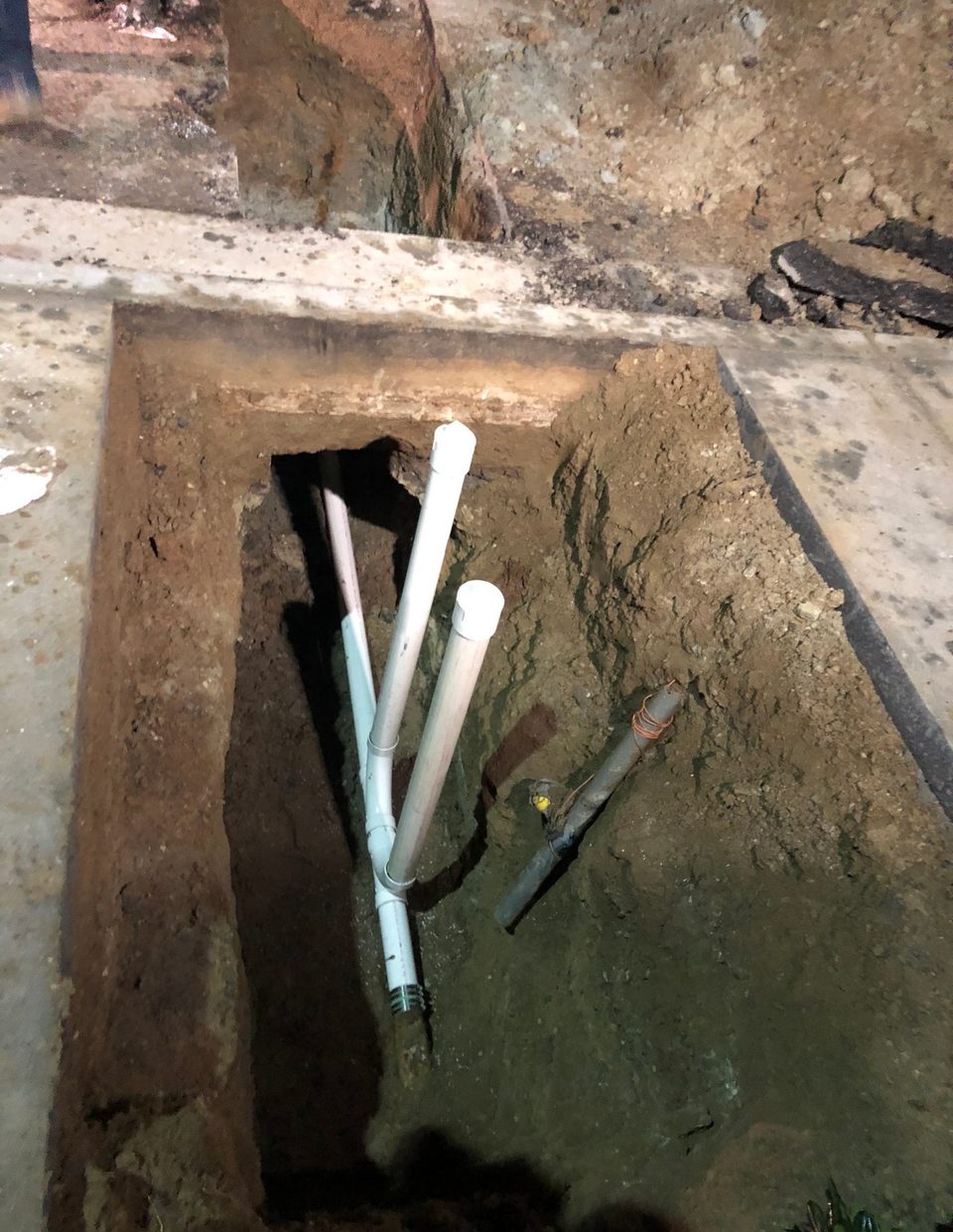 New white PVC drain lines rising from a trench cut through a concrete slab, photographed at night