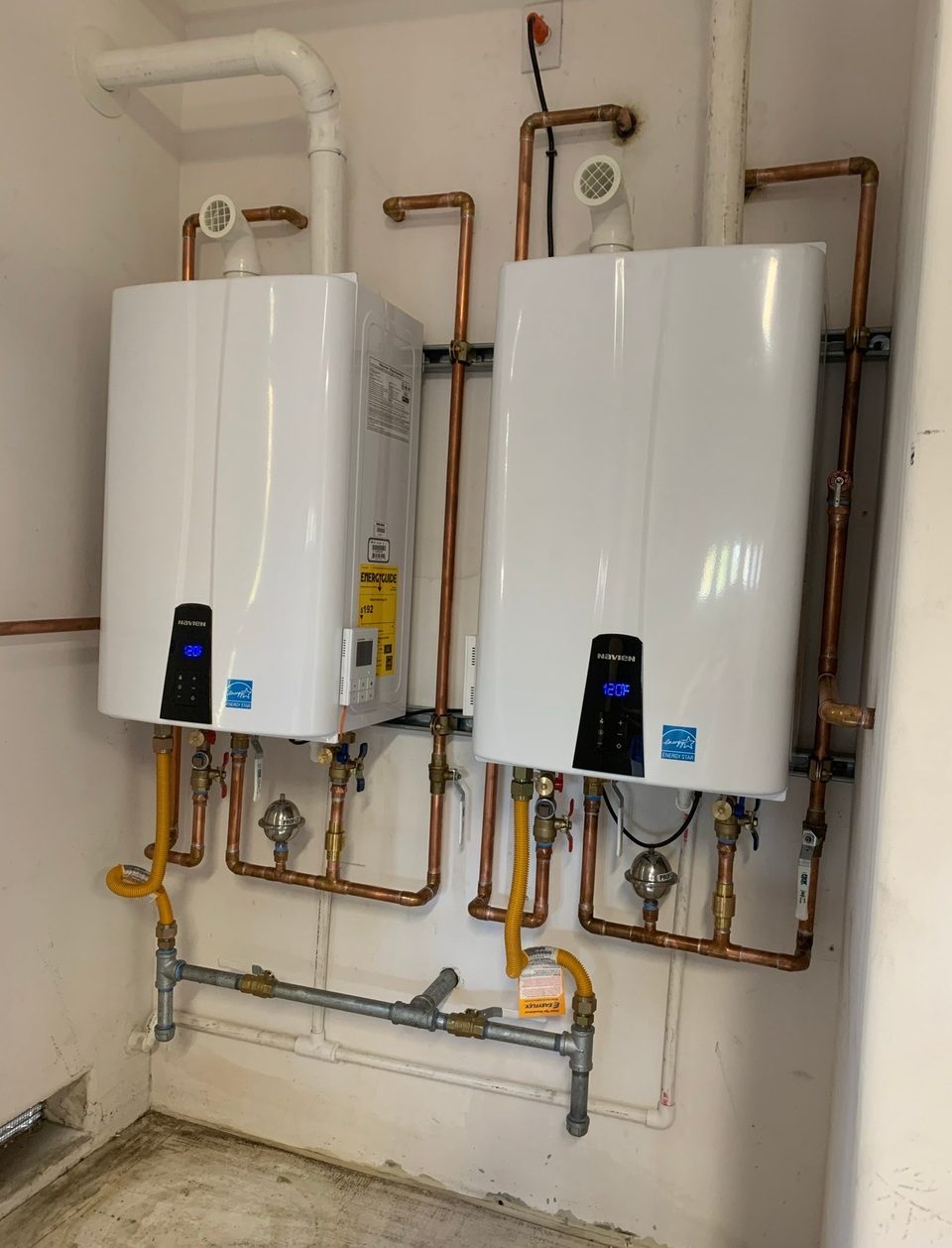 Two white Navien tankless water heaters mounted side by side on a wall, connected with copper manifolds, isolation valves and yellow gas lines